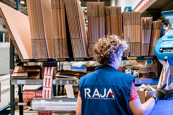 Bild eines Lagerarbeiters mit RAJA-Weste vor Regalen mit Kartonverpackungen, das die Digital- und E‑Commerce-Erfolgsgeschichte der RAJA Group veranschaulicht.